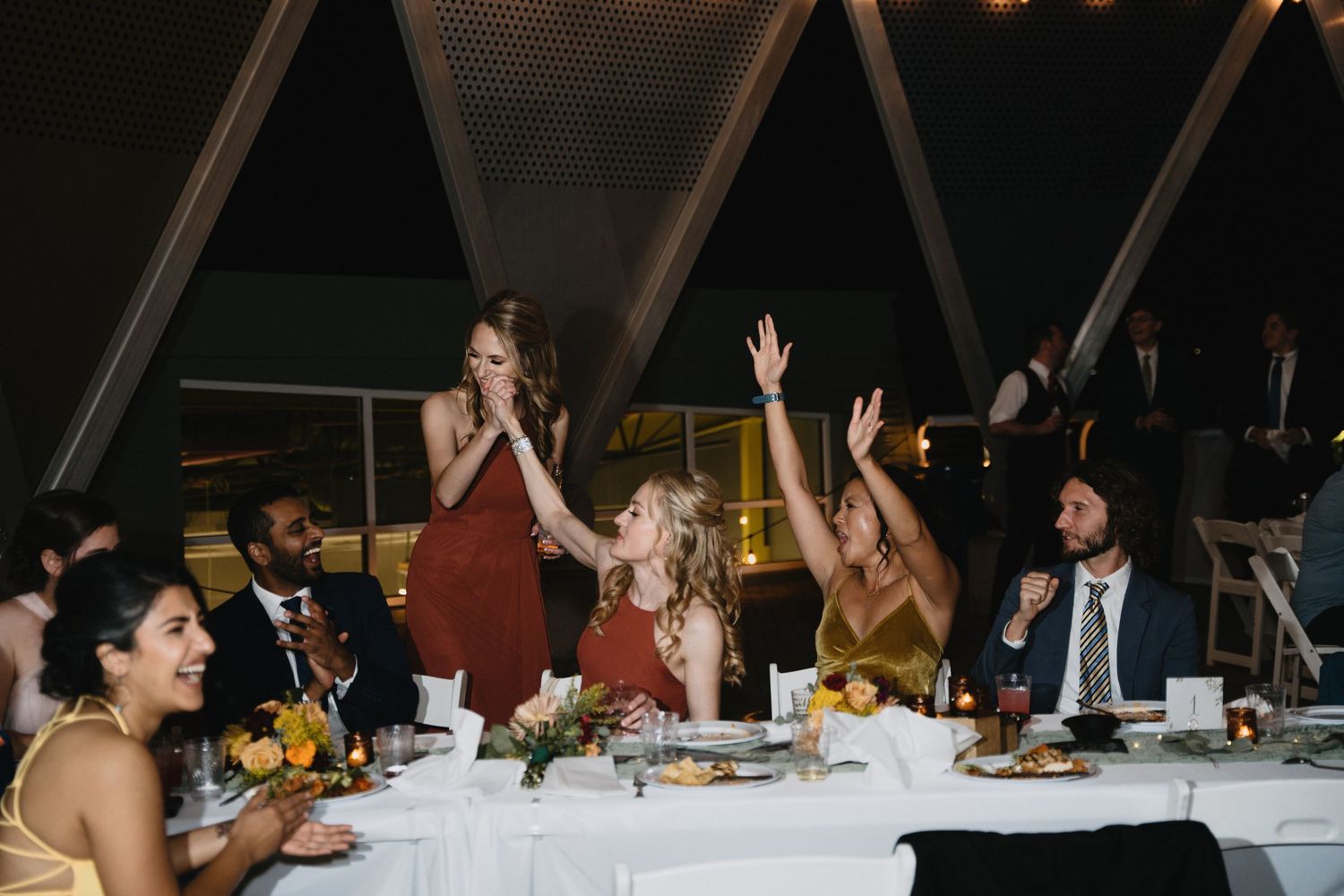 Wedding reception guests laughing and raising hands at elegantly decorated dinner table with floral centerpieces.