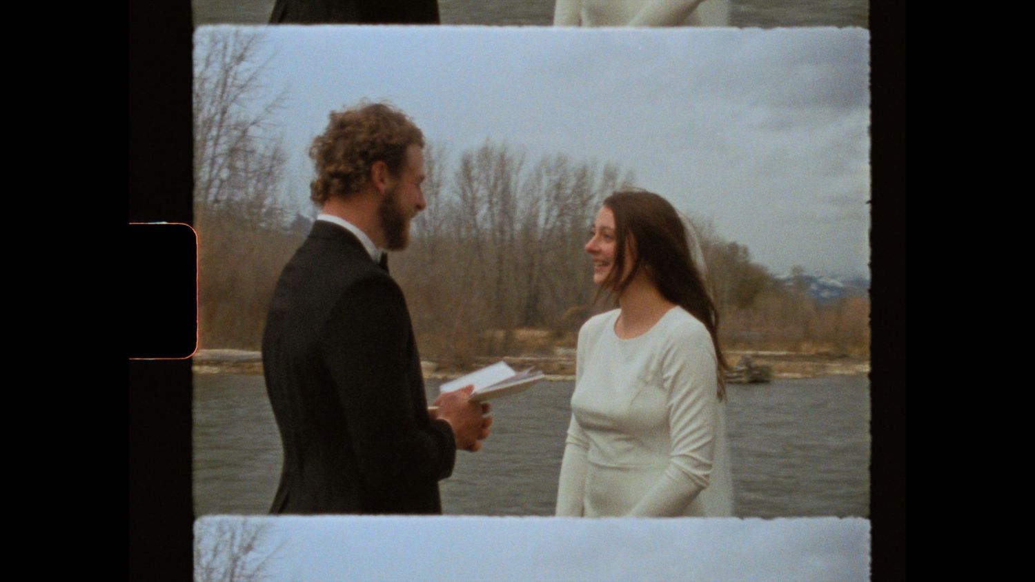A man and woman stand by a river, smiling during an outdoor ceremony, dressed in formal attire.
