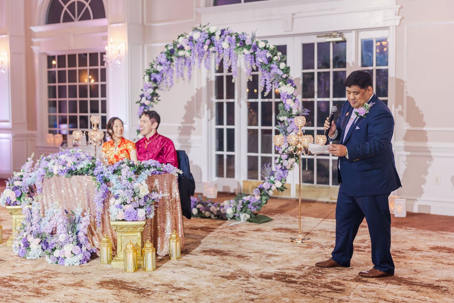 A man in a blue suit speaks at a wedding reception with a floral arch and couple seated at a decorated table.