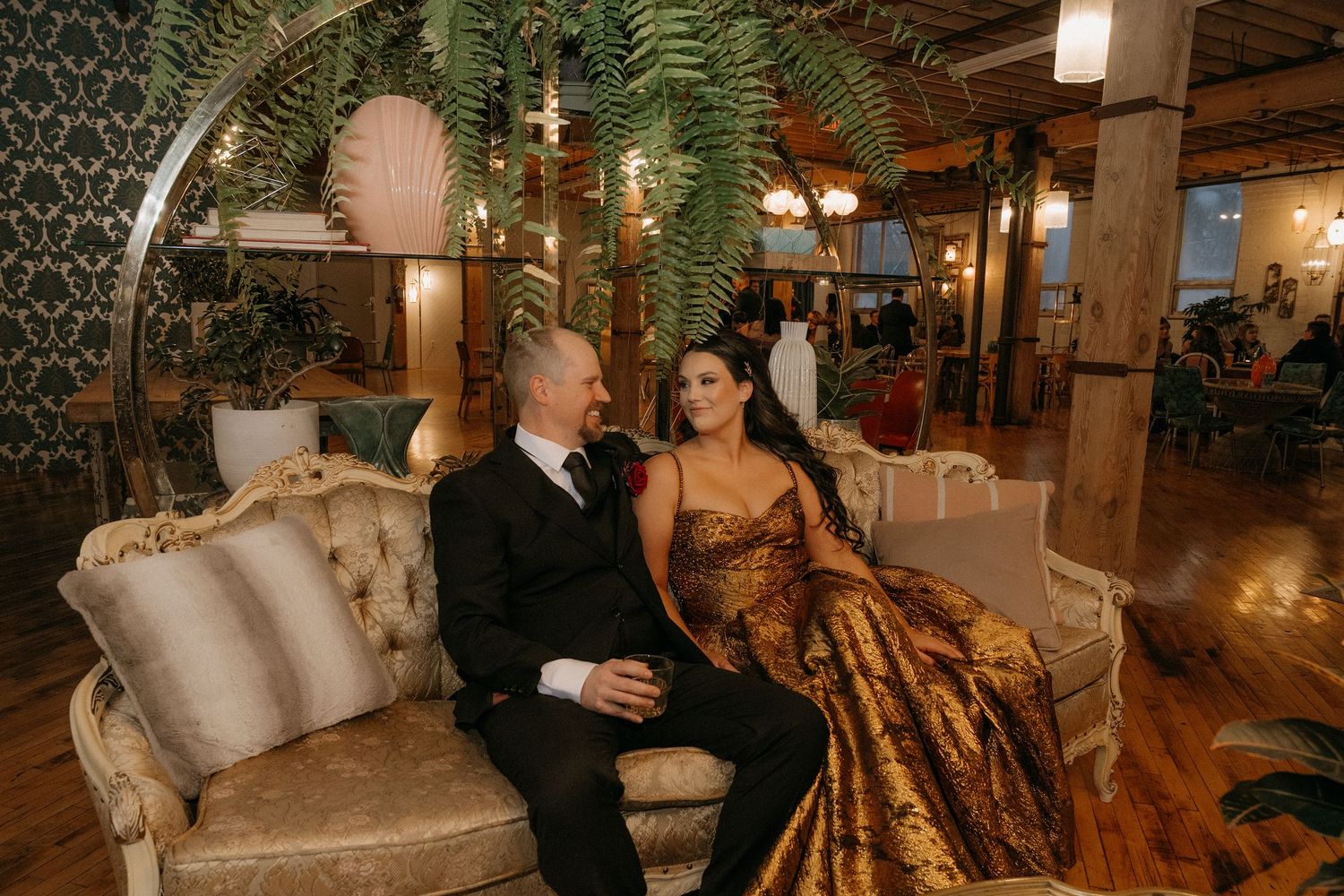 Couple in elegant formal attire sitting on vintage sofa in ornate venue with lush greenery decor.