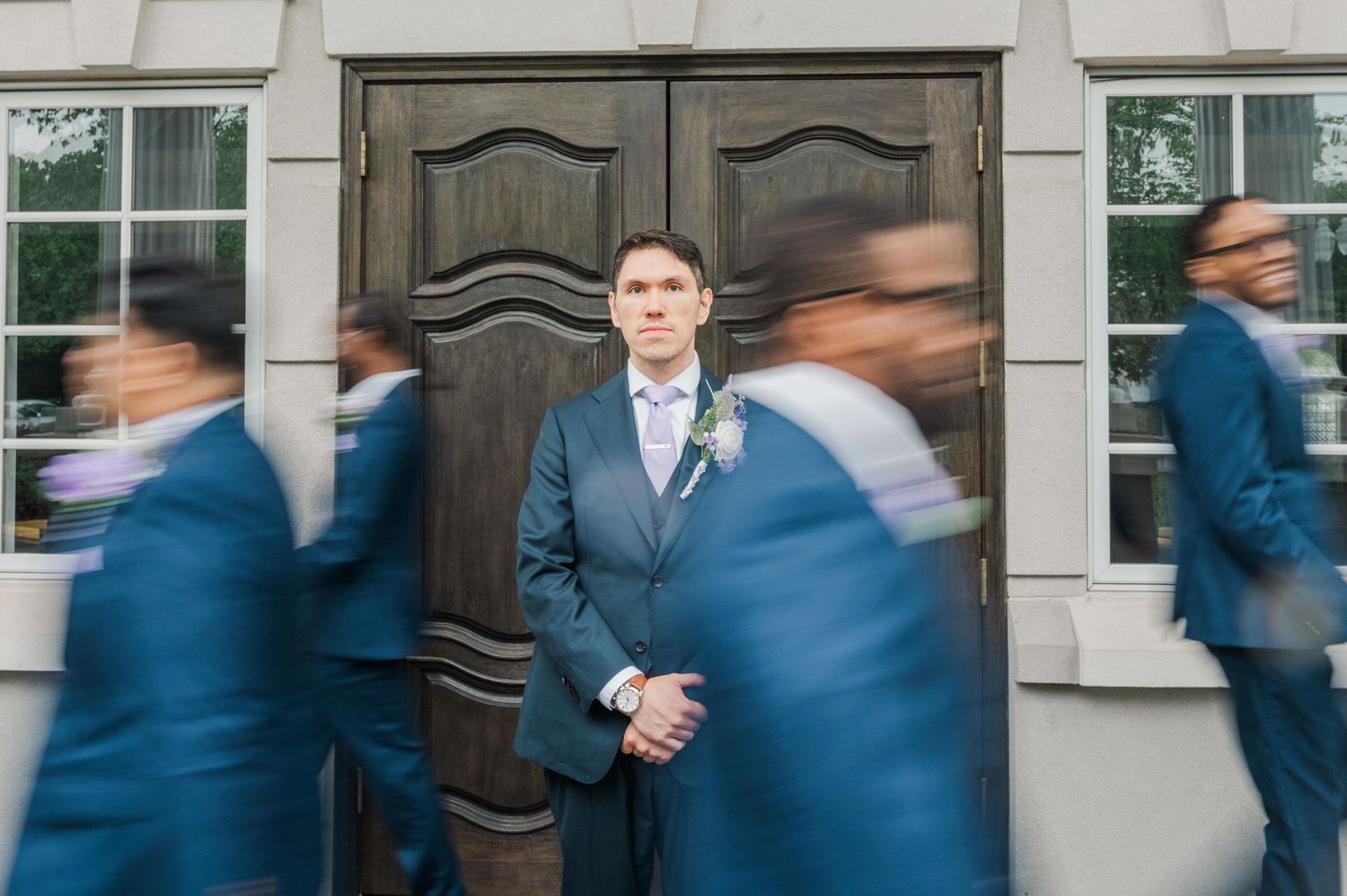 Groom stands still in blue suit before large wooden doors as blurred groomsmen in matching suits move around him.