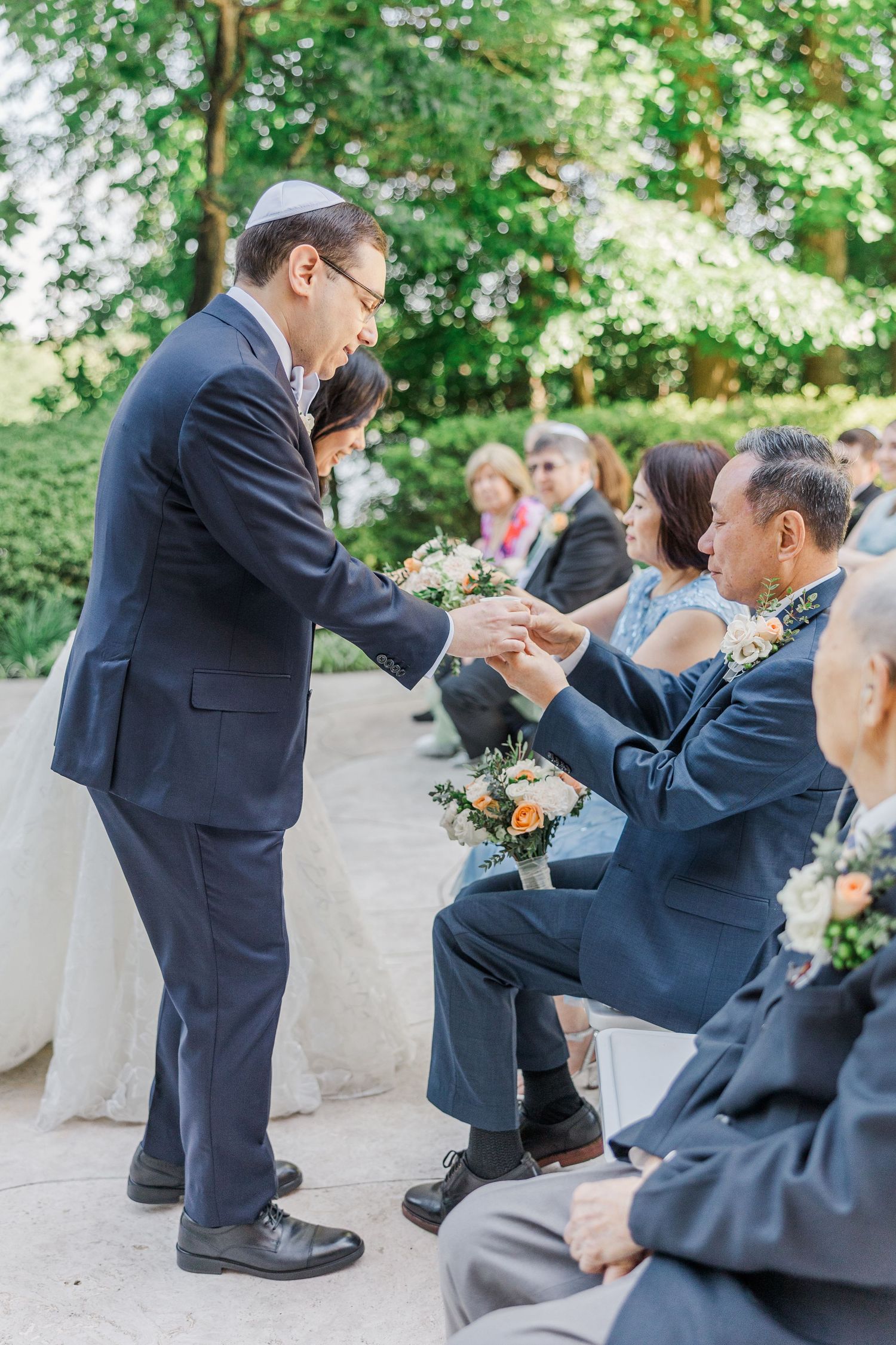 Groom in navy suit exchanges rings with seated guest during outdoor wedding ceremony surrounded by lush greenery.