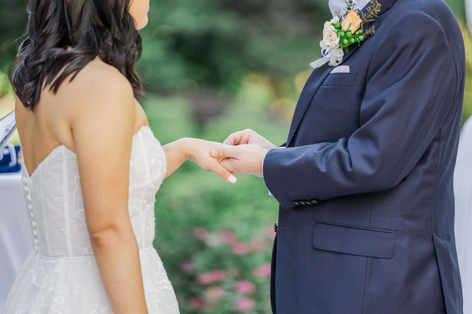 Groom placing wedding ring on bride's finger during outdoor ceremony, both dressed in elegant wedding attire.