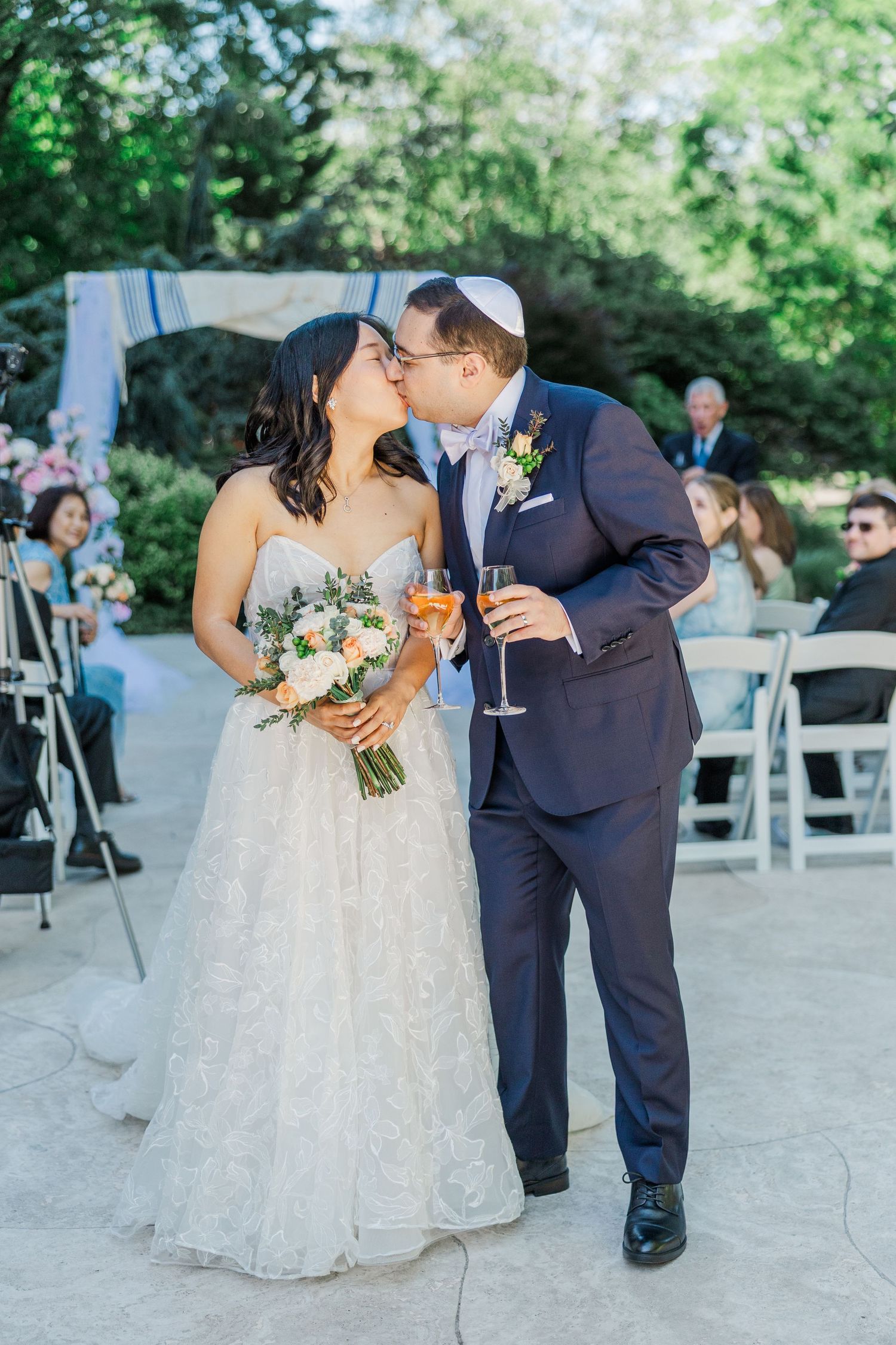 Bride and groom sharing a kiss while holding champagne glasses at an outdoor Jewish wedding ceremony.