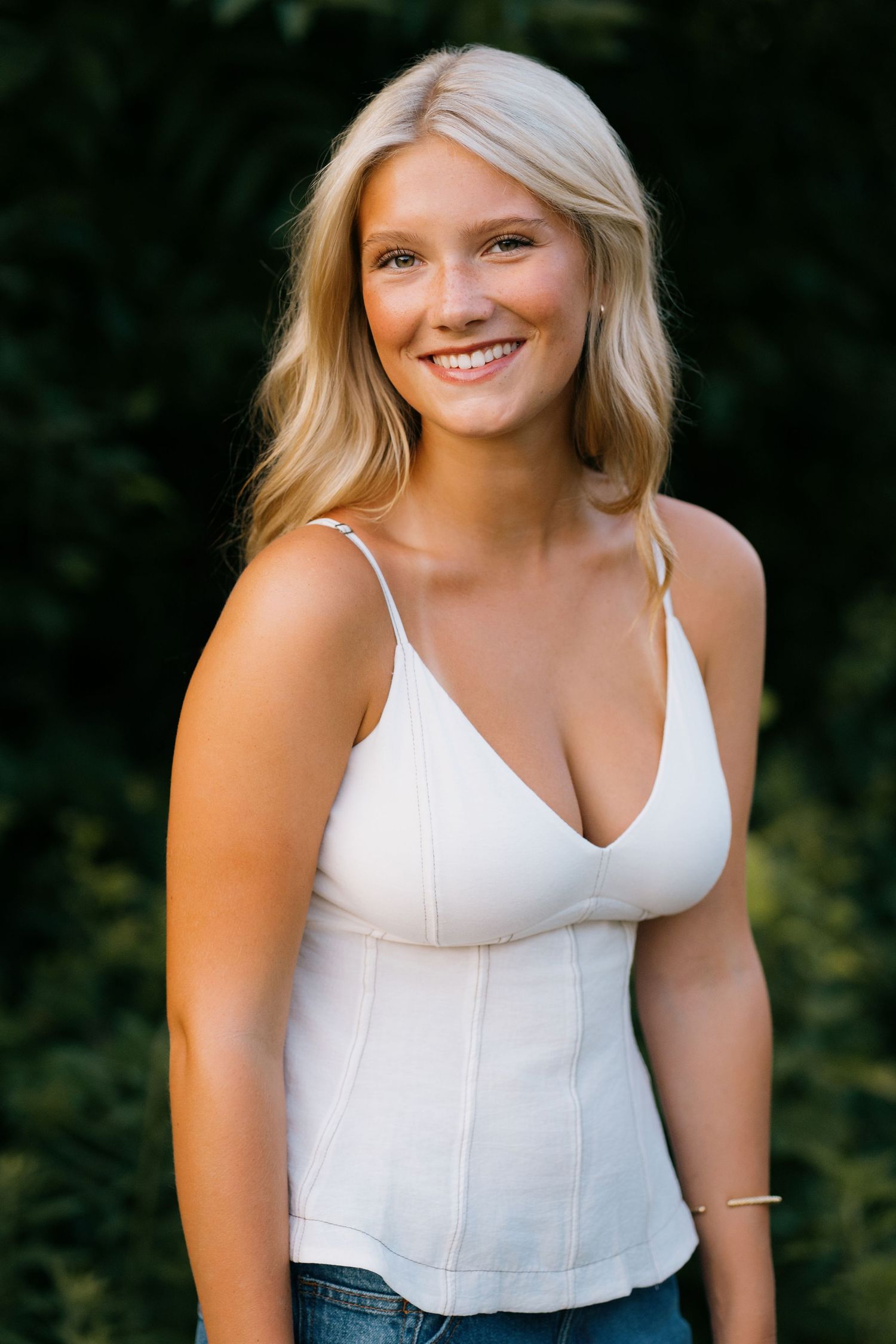 Smiling young blonde woman in white camisole top and jeans posing outdoors against green foliage backdrop.
