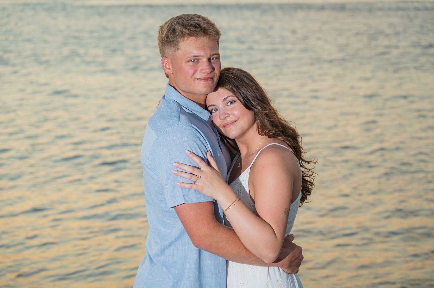Couple embracing and smiling at sunset by the water, man in blue shirt and woman in white dress.