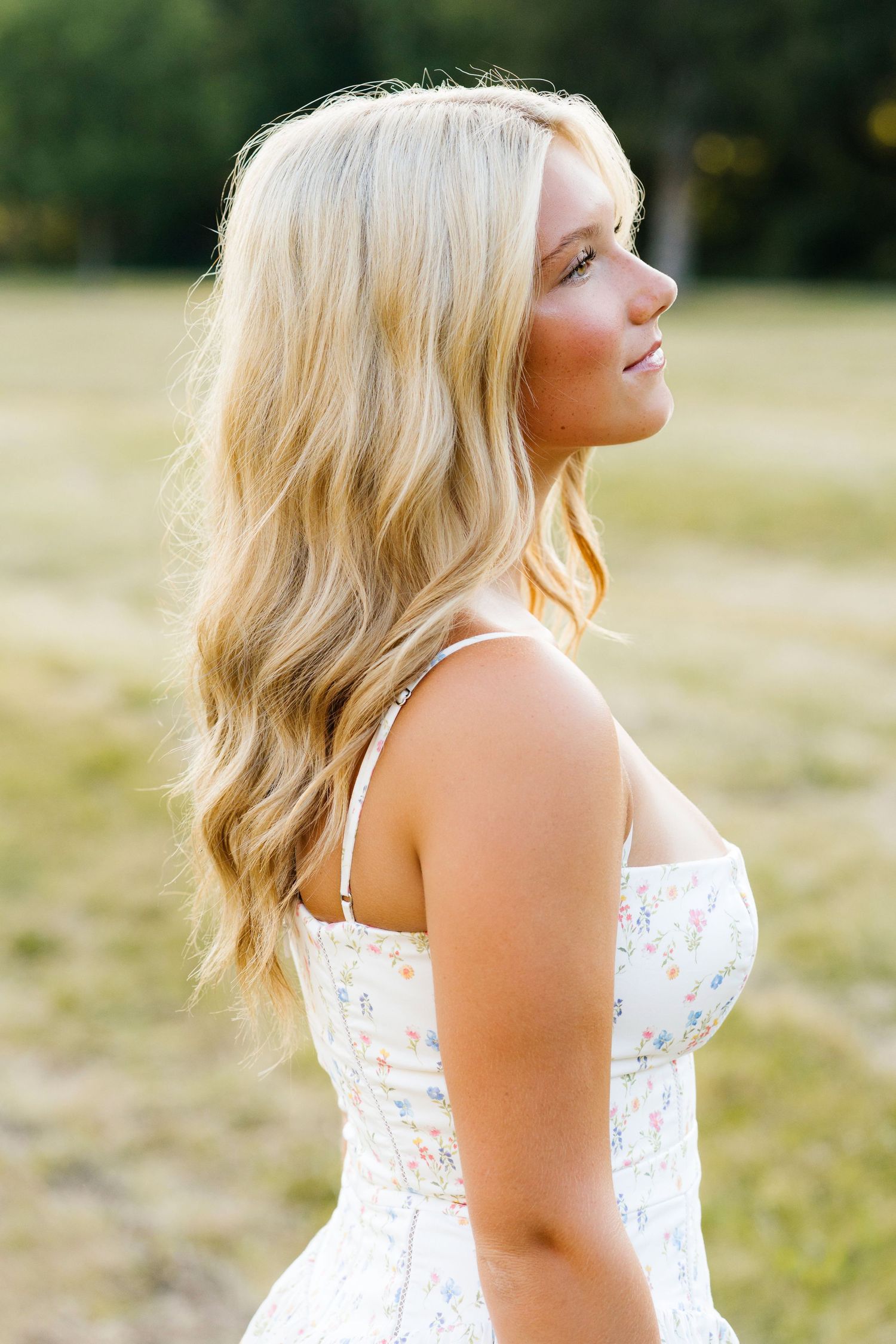 Young blonde woman in a floral sundress poses outdoors in a sunlit field, hair flowing over her shoulder.