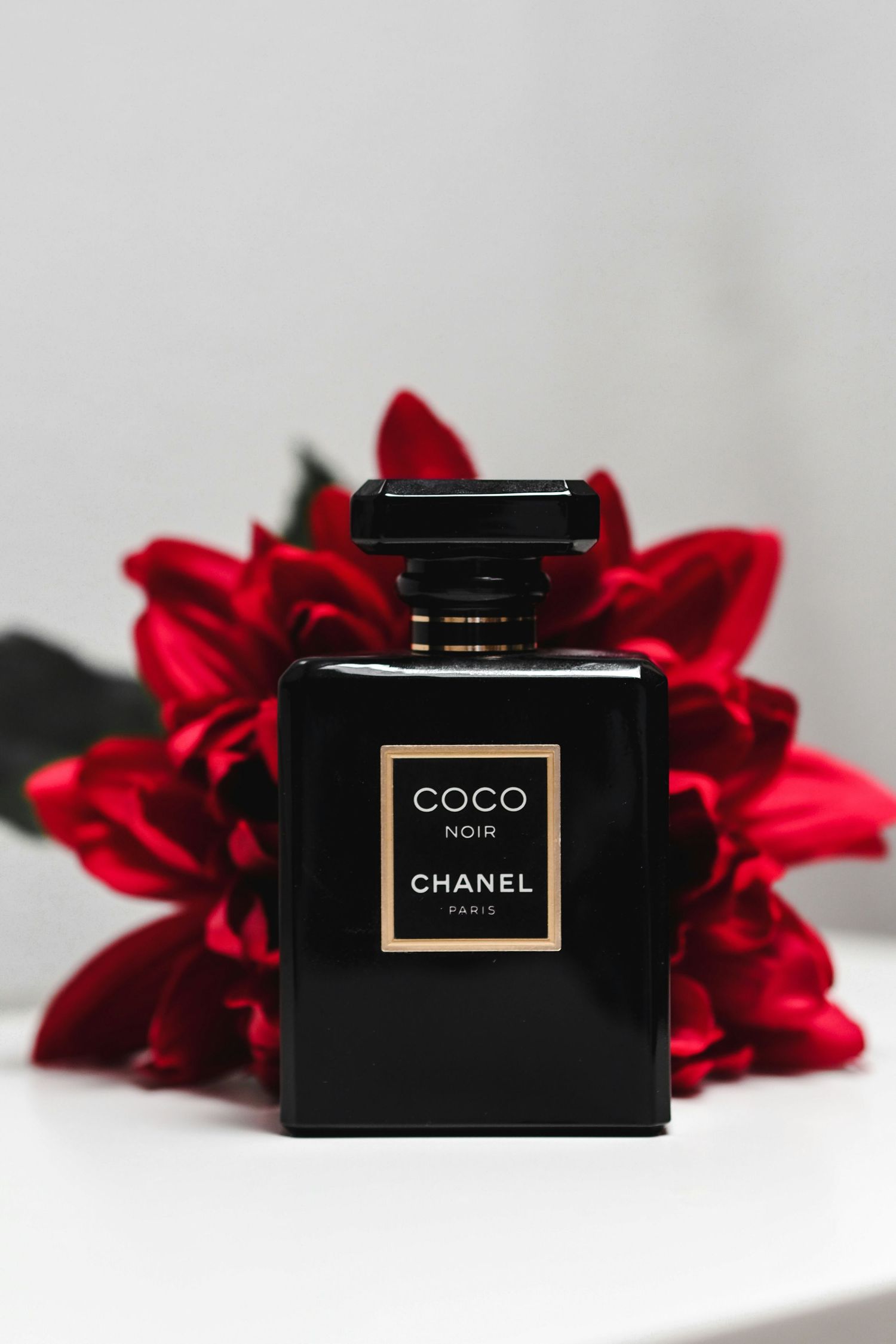 Chanel Coco Noir black perfume bottle with gold label displayed against vibrant red flowers on white surface.