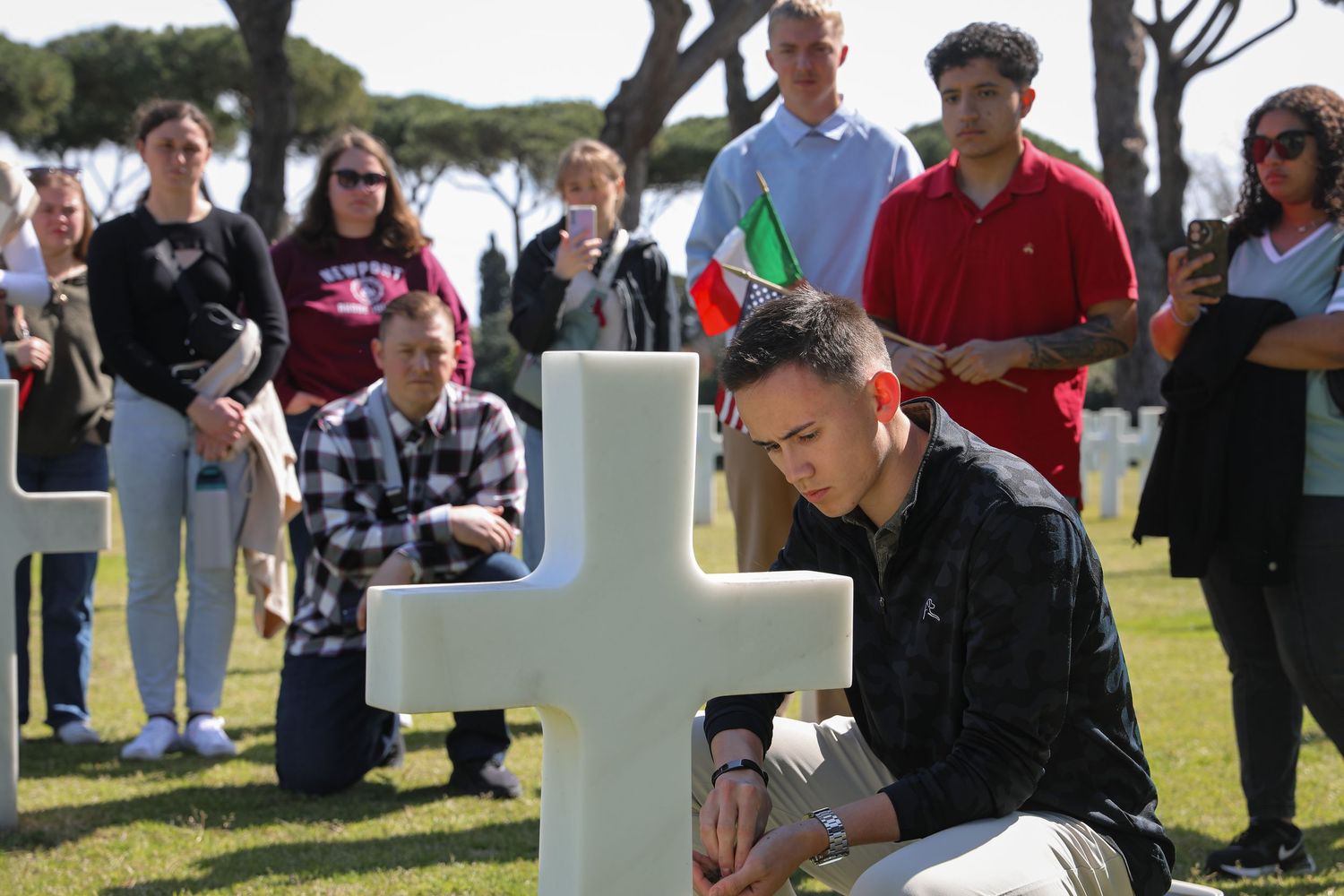 A man kneels solemnly at a white cross in a military cemetery as a crowd of visitors stands respectfully behind him.
