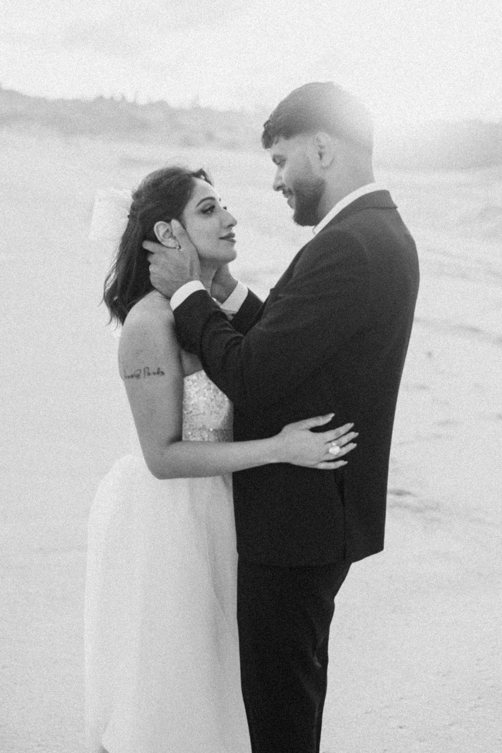 Black and white beach wedding photo of bride and groom sharing a tender moment, groom gently touching bride's face.