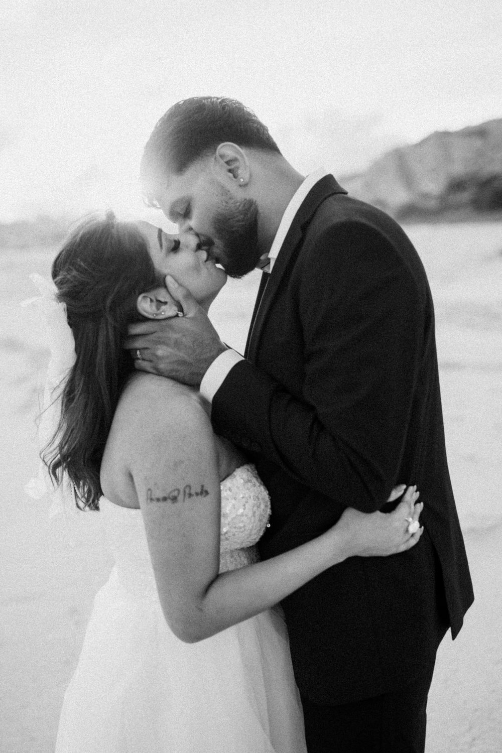 Black and white photo of a couple sharing a romantic kiss on the beach in wedding attire.