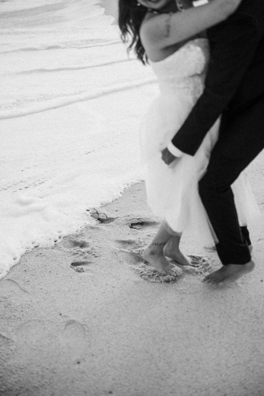 Black and white photo of a bride and groom walking barefoot on a sandy beach, leaving footprints in the sand.