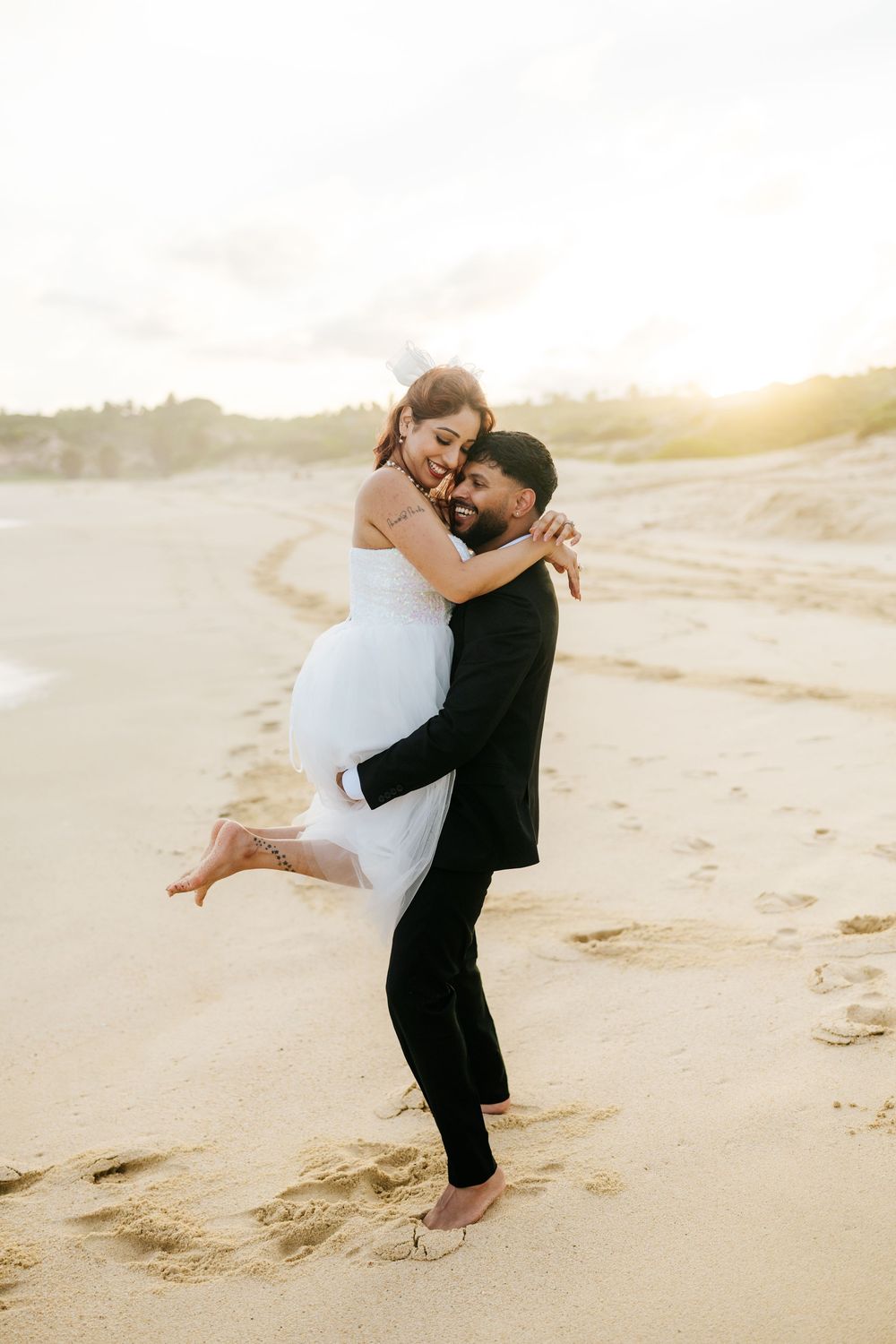 Groom in black suit lifts smiling bride in white dress on sandy beach at golden sunset.