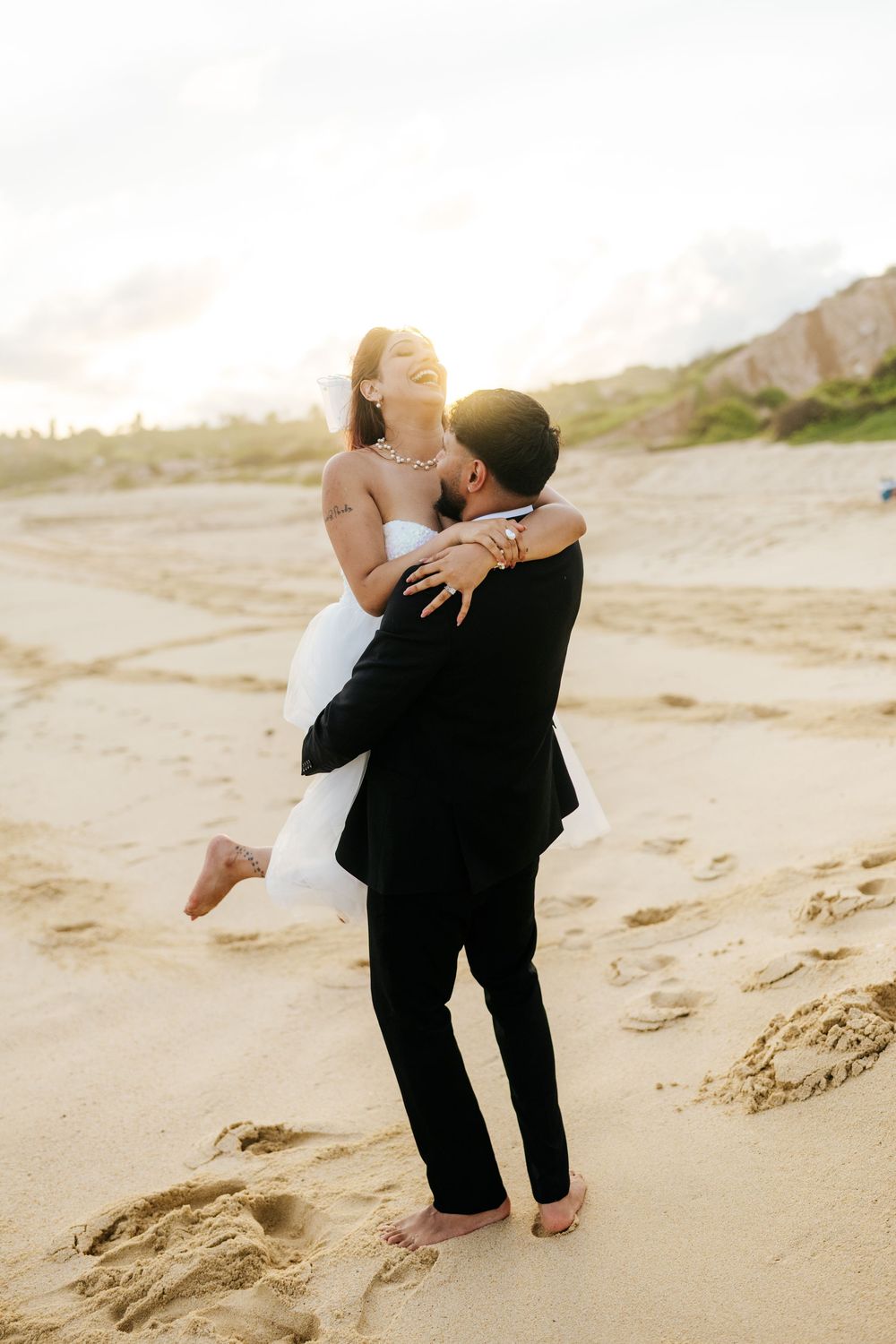 Groom lifting bride in white dress on sandy beach at sunset, sharing a joyful romantic moment.