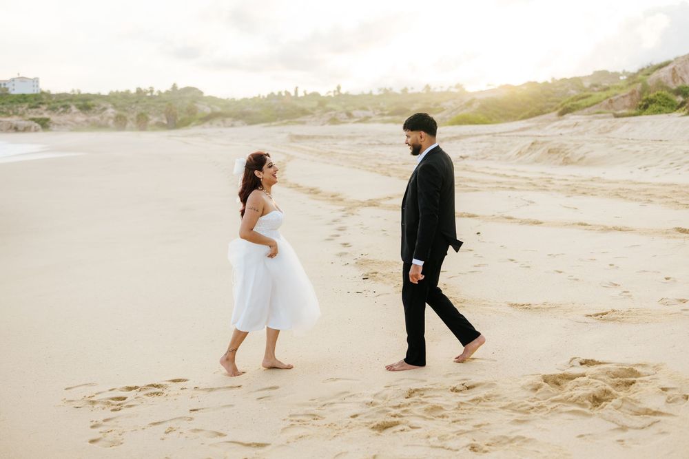 Couple in wedding attire walking barefoot on sandy beach, bride in white dress and groom in black suit.