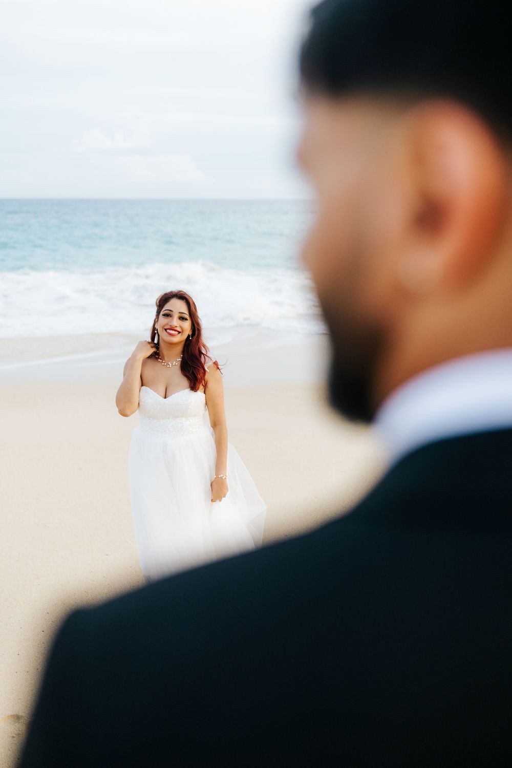 Smiling bride in white gown stands on sandy beach as groom in black suit looks on, with ocean waves in background.