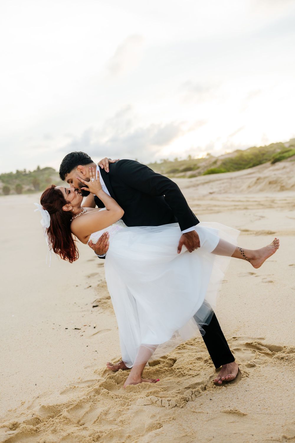 Groom dips bride for a romantic kiss on sandy beach at sunset, bride in white gown, groom in black suit.