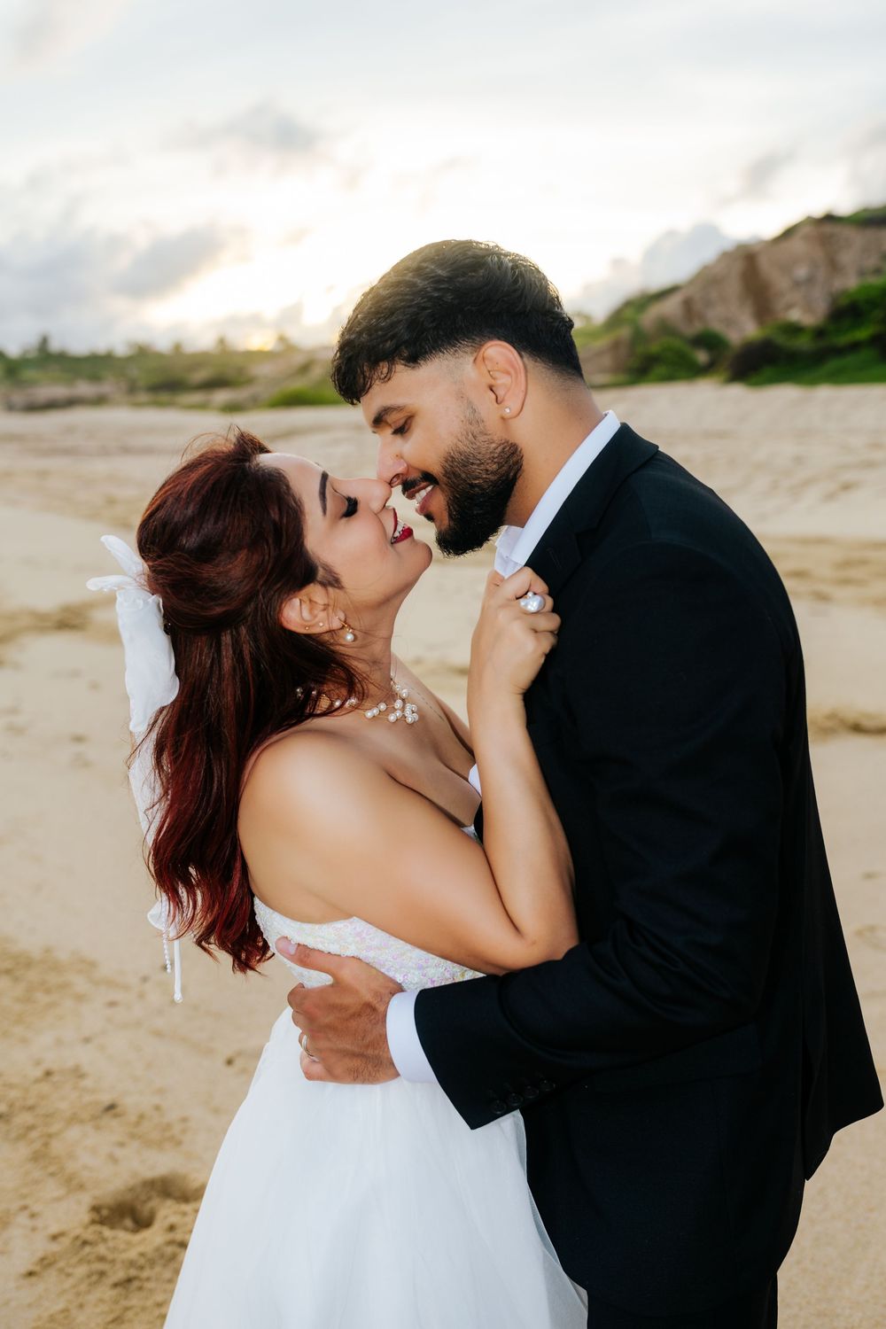 A bride and groom share a romantic moment on a sandy beach, with the bride in a white gown and groom in a black suit.