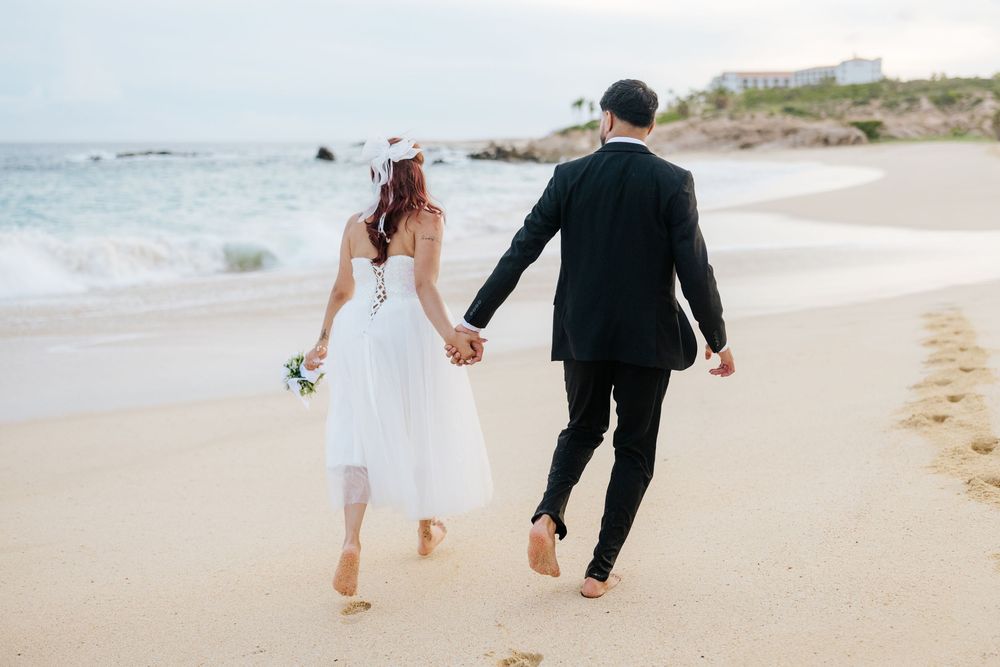 Newlywed couple walking hand-in-hand barefoot on a sandy beach, bride in white dress holding bouquet, groom in black suit.