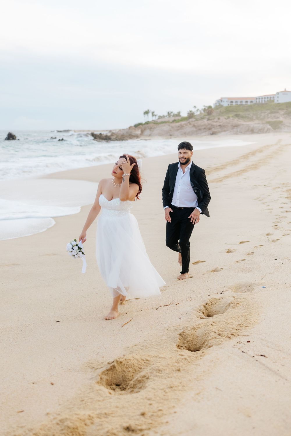Bride and groom walking barefoot on sandy beach, woman in white dress holding bouquet, man in black suit smiling.