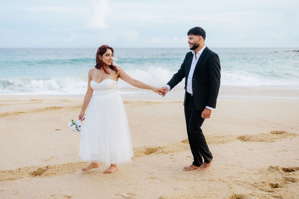 Bride and groom holding hands on a sandy beach, waves in background, bride in white dress holding bouquet.