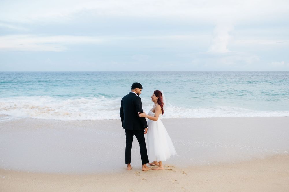 Couple in wedding attire holding hands on a sandy beach with ocean waves and cloudy sky backdrop.