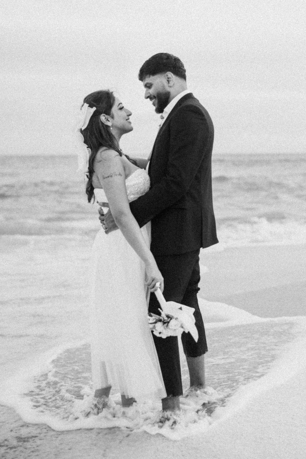 Black and white photo of a bride and groom standing in ocean waves on a beach, smiling at each other on their wedding day.