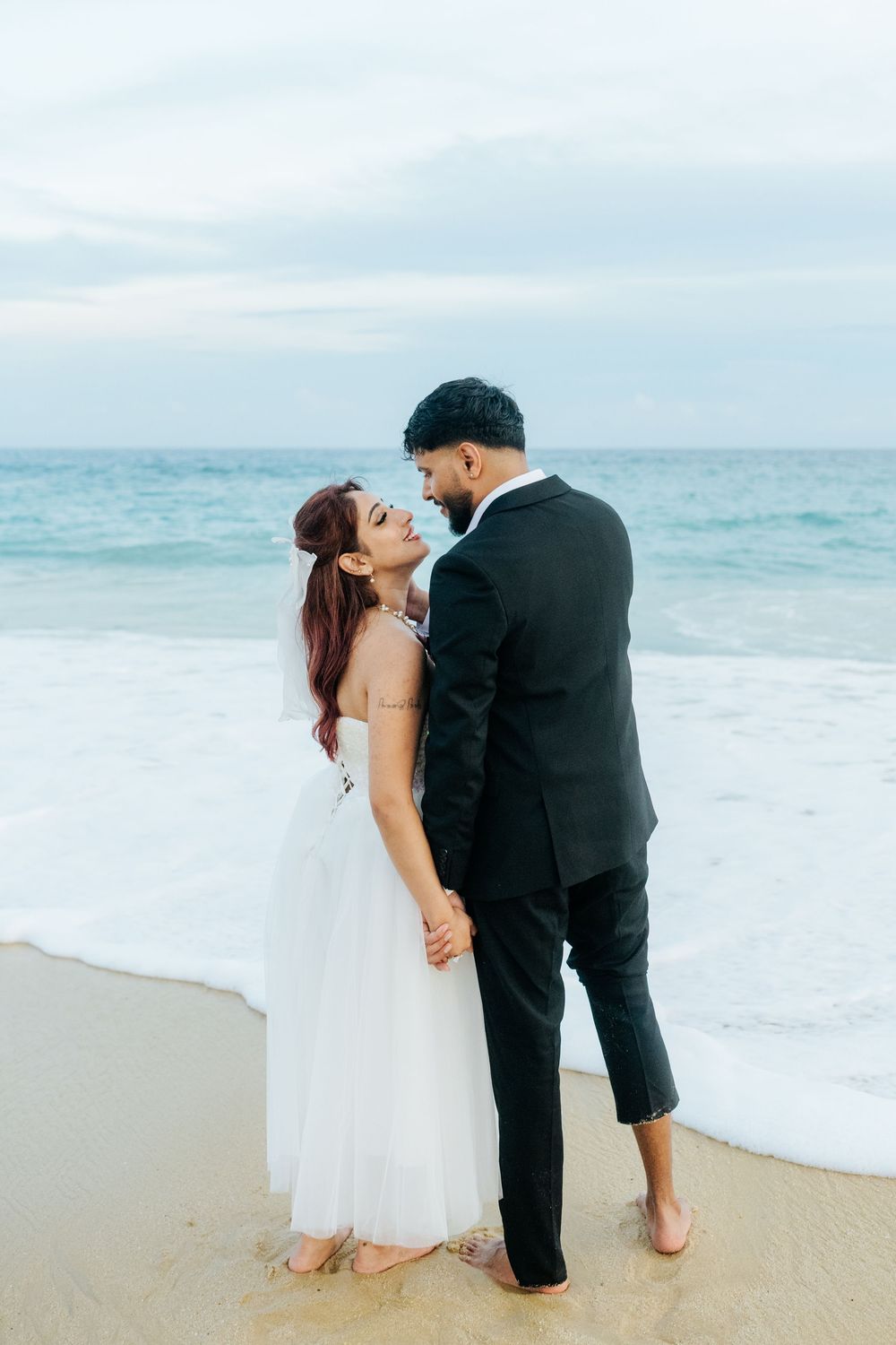 A couple in wedding attire shares a romantic moment on a beach, with the bride in white and groom in a black suit.