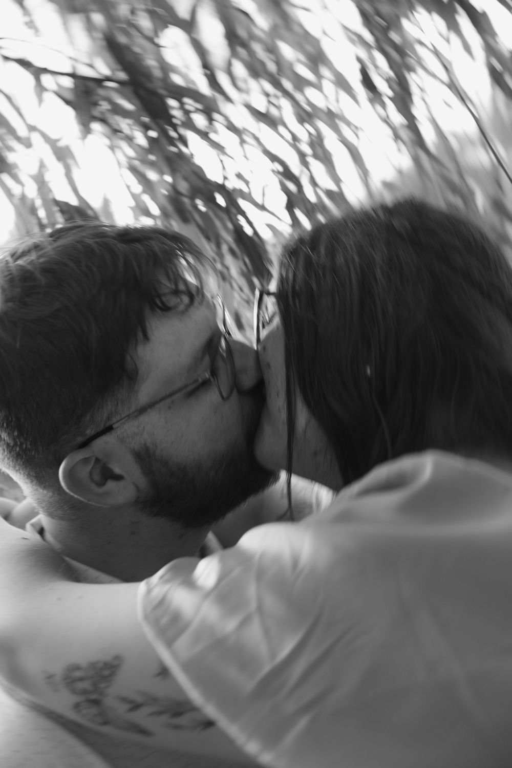 Black and white photo of a couple sharing a tender kiss under leafy branches outdoors.