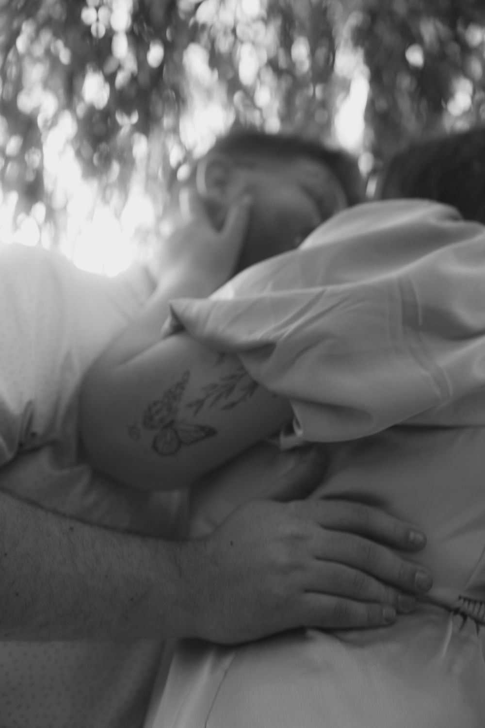 Black and white photo of two people sharing a warm embrace outdoors, with a butterfly tattoo visible on one person's hand.