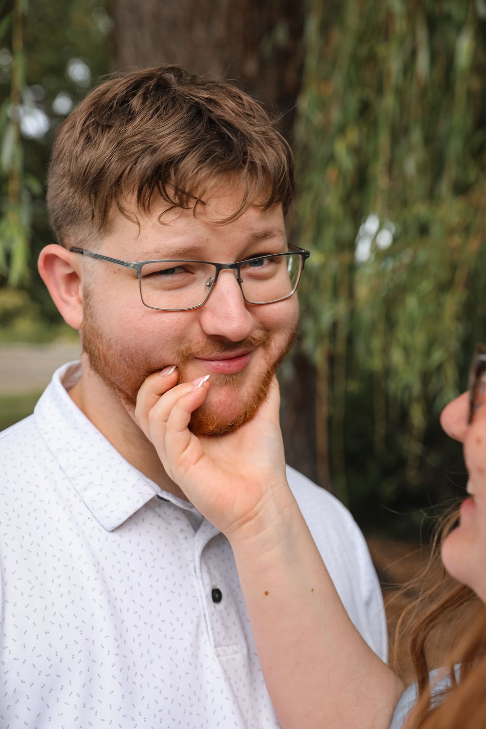 Young man with glasses and red beard smiling as woman touches his face outdoors.