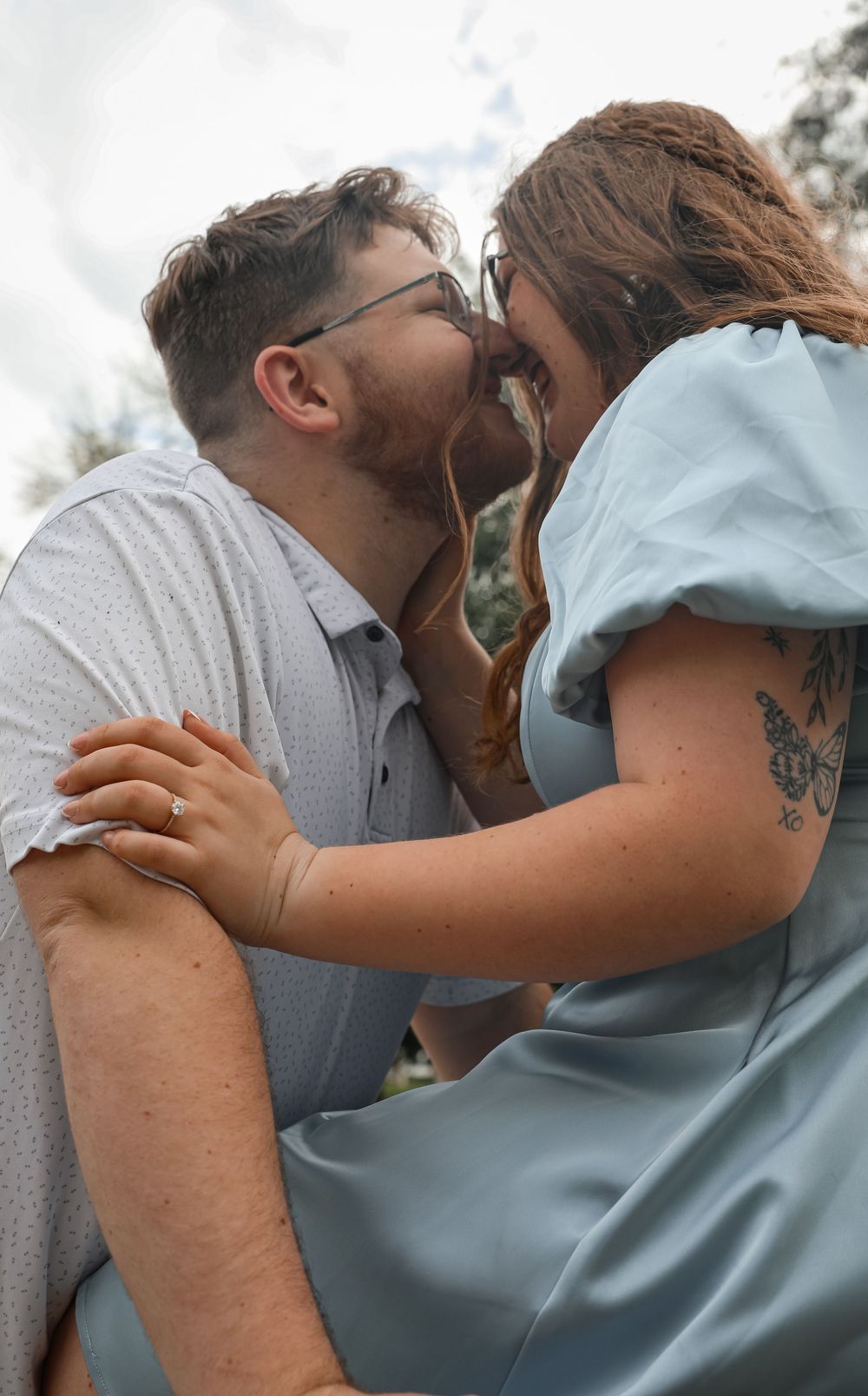 A couple shares a romantic kiss outdoors, the woman in a light blue dress with a butterfly tattoo embracing a man in a gray shirt.