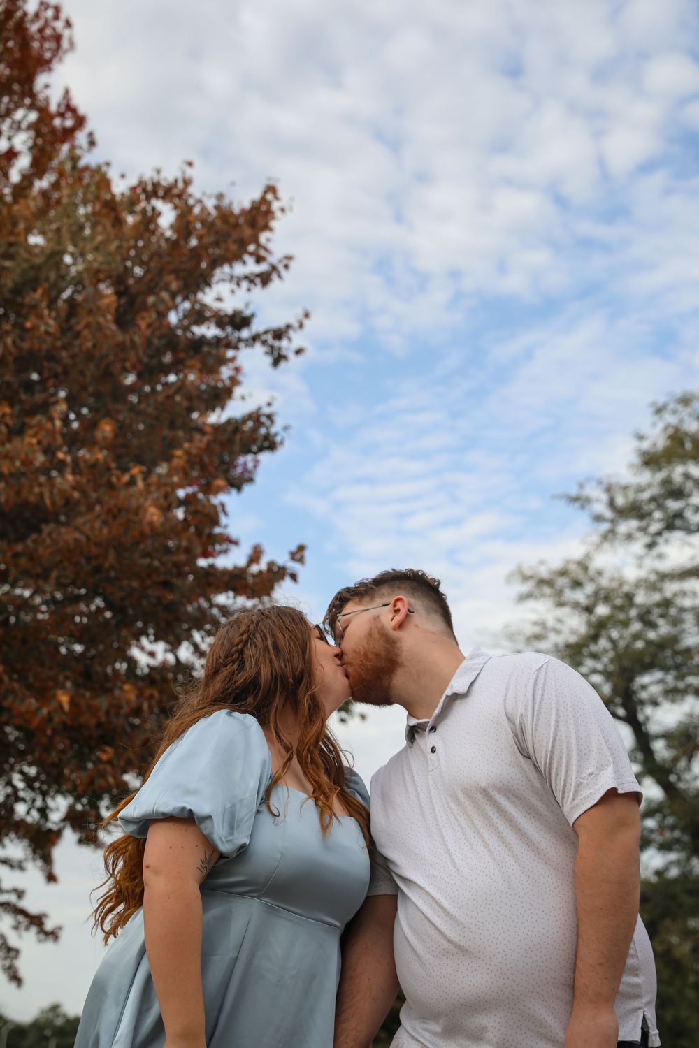 Couple sharing a romantic kiss outdoors with autumn trees and cloudy blue sky in the background.