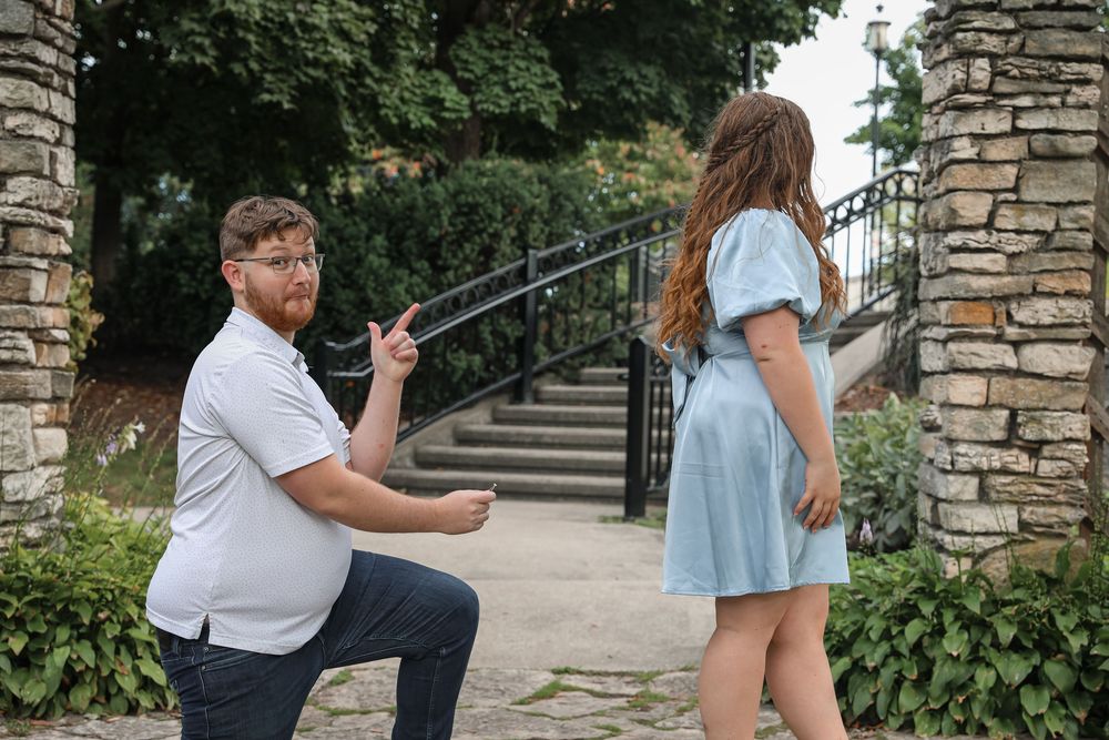 Man proposing on one knee to woman in blue dress near stone archway in garden.
