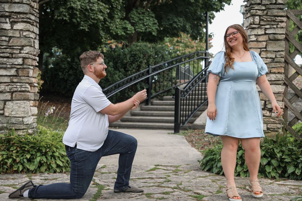 Man proposing on one knee to woman in blue dress in a garden setting.