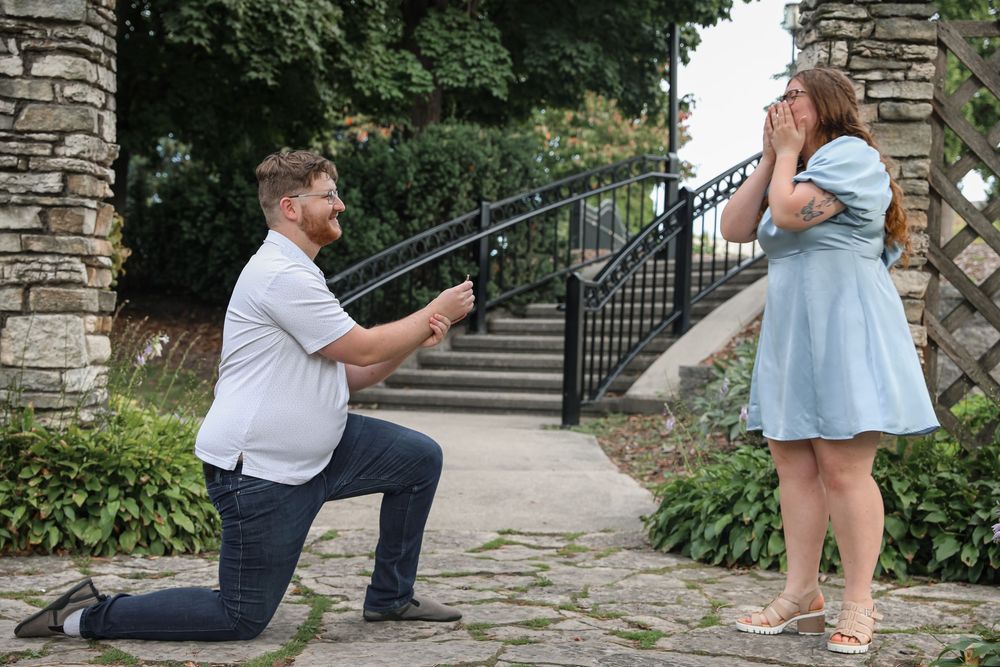 Man proposing on one knee to emotional woman in blue dress outdoors near stone steps and garden.