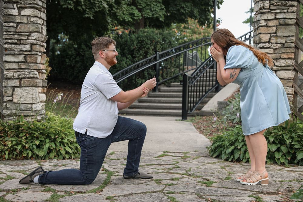 Man on one knee proposing to emotional woman in blue dress outdoors near stone steps and garden.