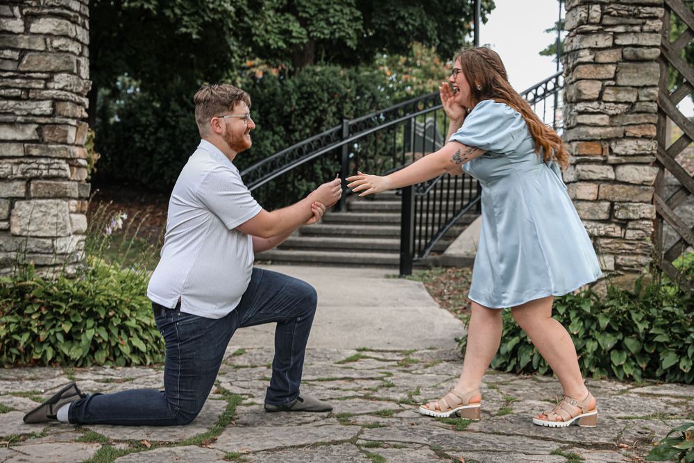 Man kneeling on one knee proposing to a woman outdoors near stone pillars and garden steps.