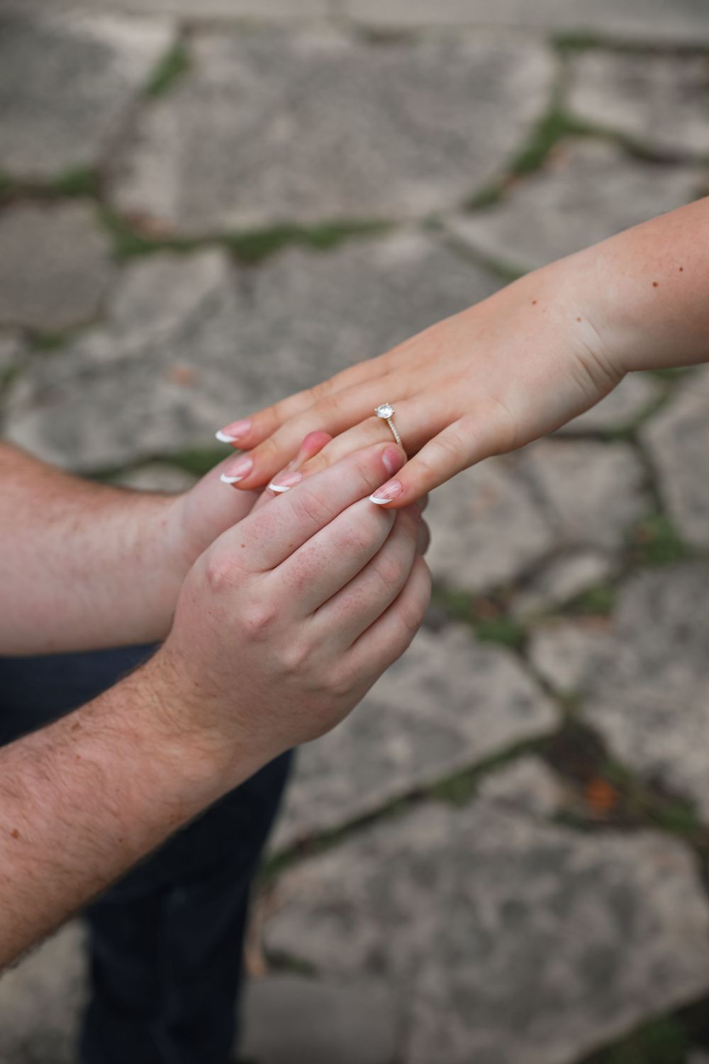Man holding woman's hand with diamond engagement ring on stone pavement background.