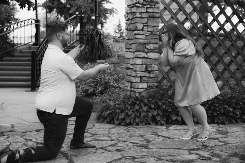 Man kneeling on one knee proposing to emotional woman in garden, black and white engagement photo.