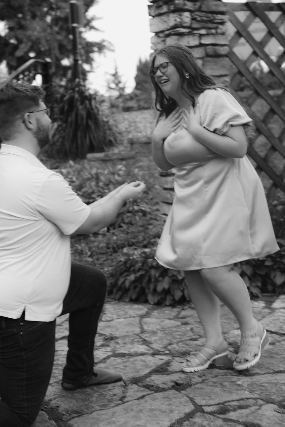 Man on one knee proposing to surprised woman in white dress in a garden setting, black and white photo.