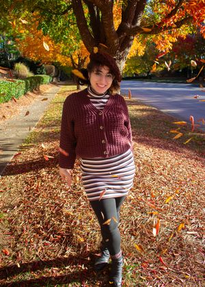Woman in burgundy sweater and striped skirt smiling outdoors among falling autumn leaves.