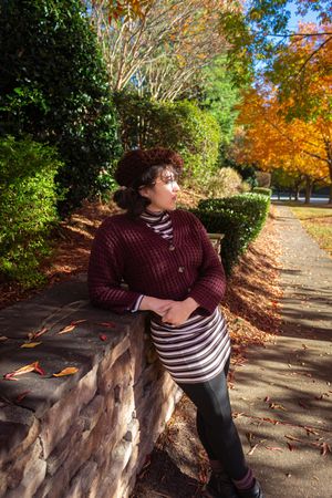Woman in burgundy cardigan and striped skirt leaning against stone wall in colorful autumn park setting.