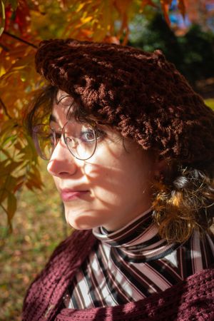 Young woman in brown crochet beret and glasses poses outdoors among vibrant autumn foliage in fall.