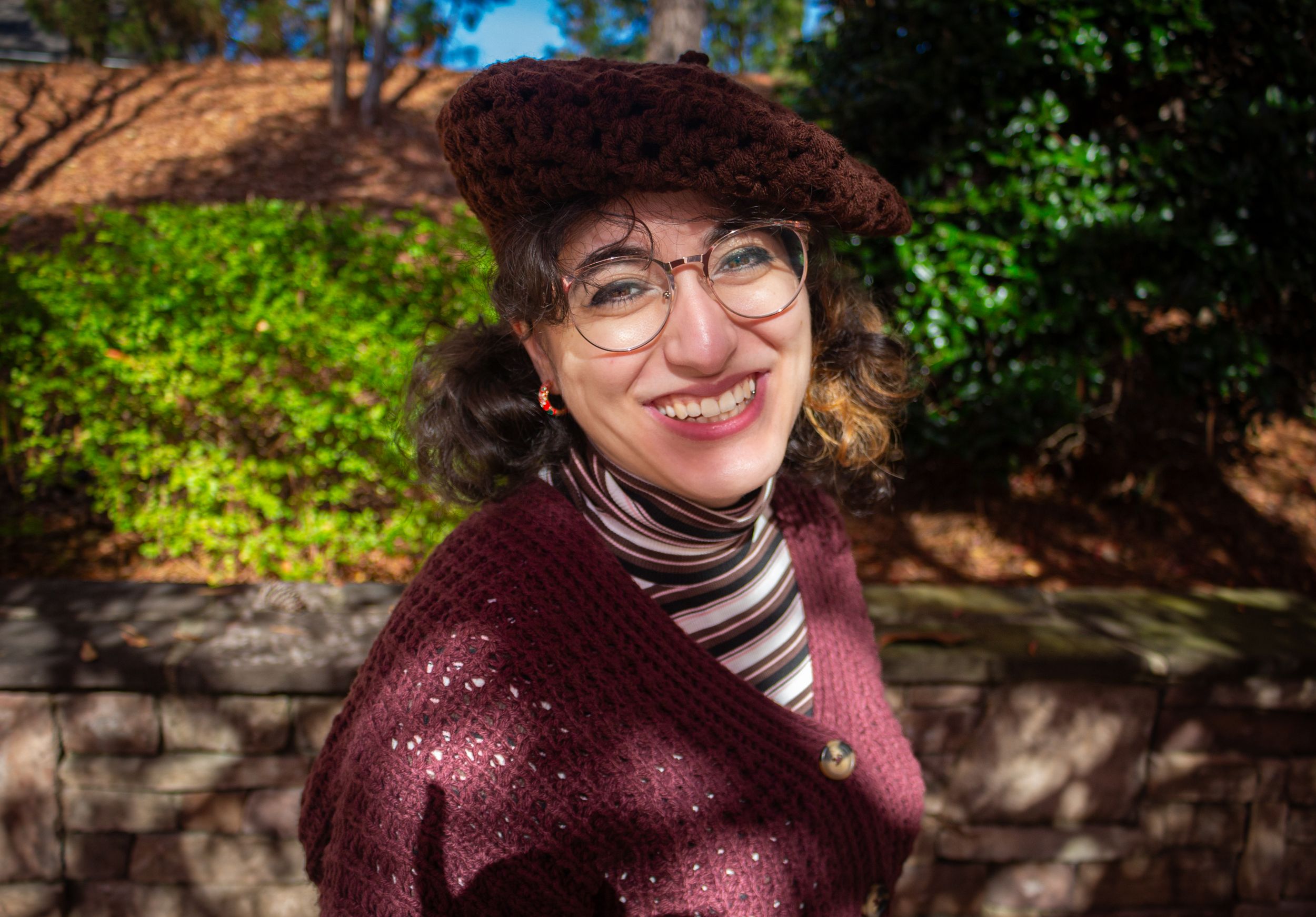 Smiling woman wearing glasses and a dark beret hat outdoors in a garden setting.