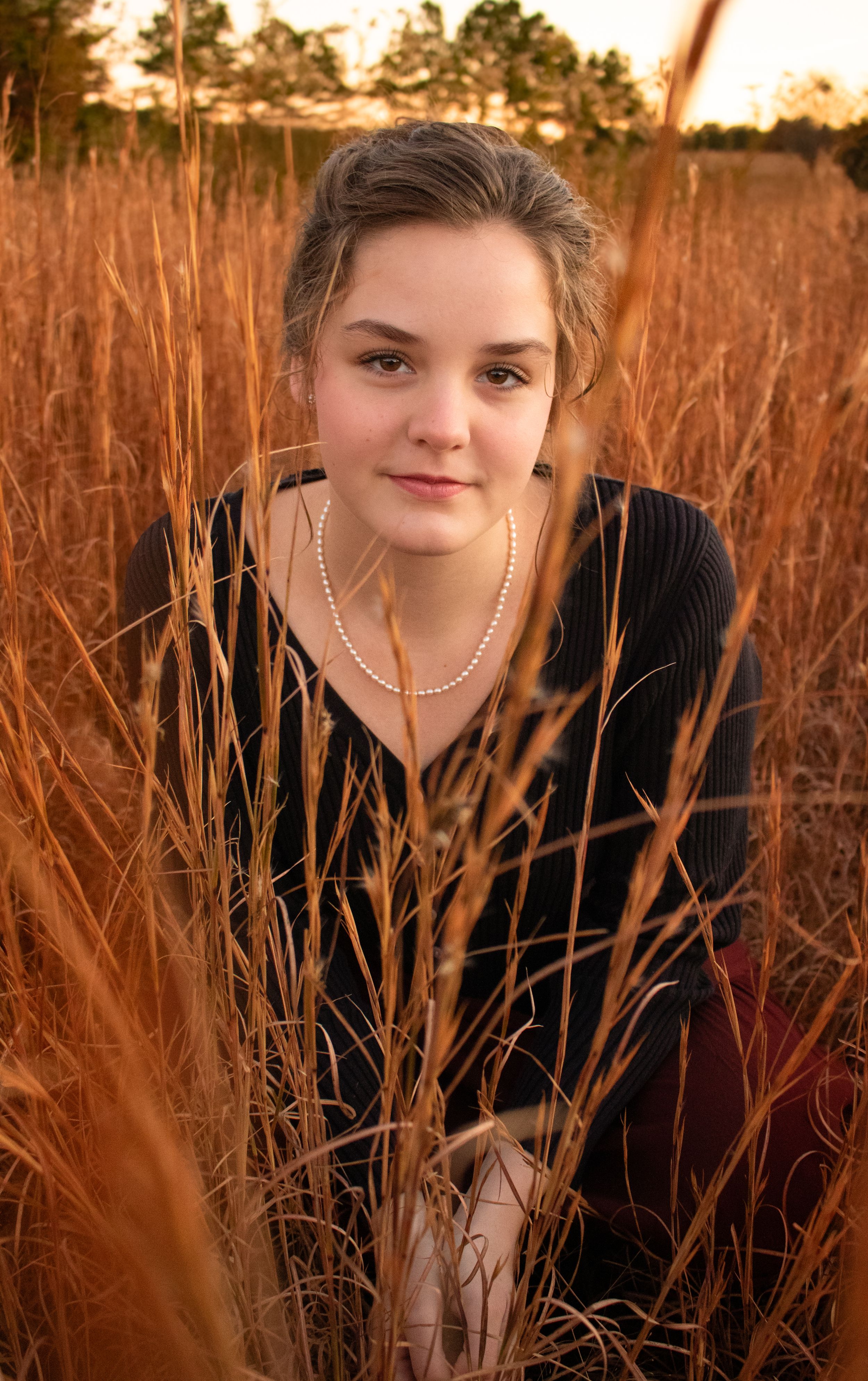 Young woman in black top and pearl necklace sitting in golden tall grass field during sunset.