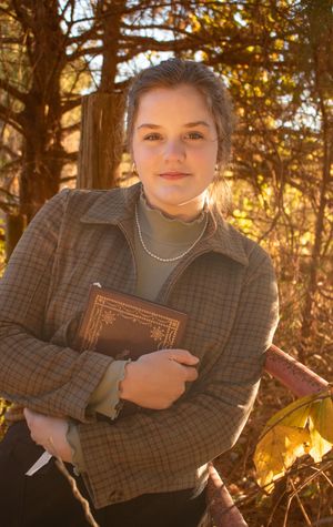 Young woman holding a vintage book outdoors in autumn sunlight, wearing a plaid jacket and pearl necklace.