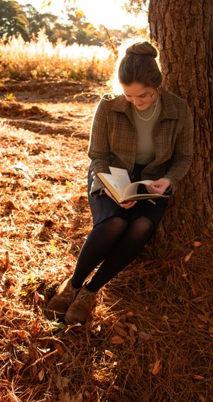 A young woman in a plaid jacket reads a book leaning against a tree surrounded by autumn leaves in golden sunlight.