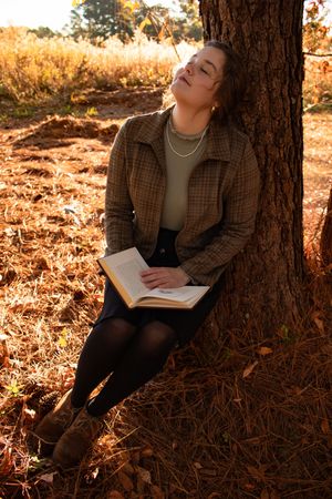 Young woman in plaid jacket sits against autumn tree, eyes closed, holding open book in golden fall light.