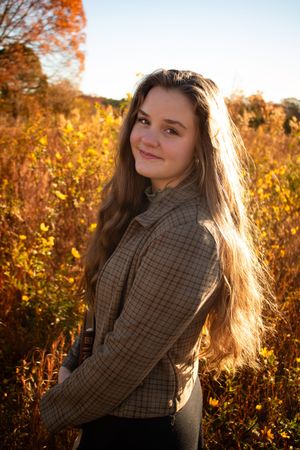 Young woman with long hair in plaid jacket smiling over shoulder in golden autumn field at sunset.